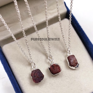 May include: Three silver necklaces, each featuring a raw, dark red gemstone pendant. The pendants have a natural, rough texture and are set in silver bezels. The necklaces are presented in a blue jewellery box. The text "PURESOULJEWELS" is visible.