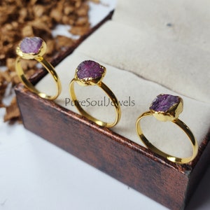 Può includere: Tre anelli d'oro con pietre preziose viola. Gli anelli sono esposti in una scatola di velluto marrone. PureSoulJewels.