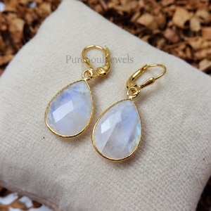 May include: A pair of gold-toned leverback earrings featuring faceted, pear-shaped moonstone gems with a translucent, iridescent white appearance. The earrings are set in a simple gold metal frame, showcasing a clean, drop-style design.