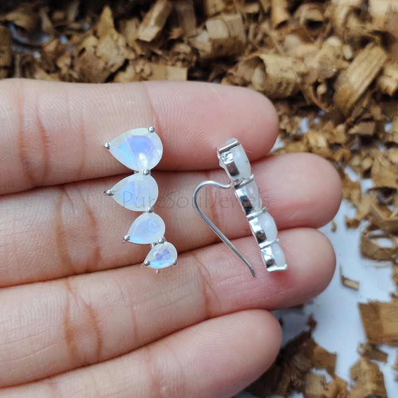 May include: A pair of silver ear climbers featuring four pear-shaped moonstones arranged in a graduated vertical line. Each stone is set in a silver prong setting, with a curved wire backing designed to follow the contour of the earlobe.