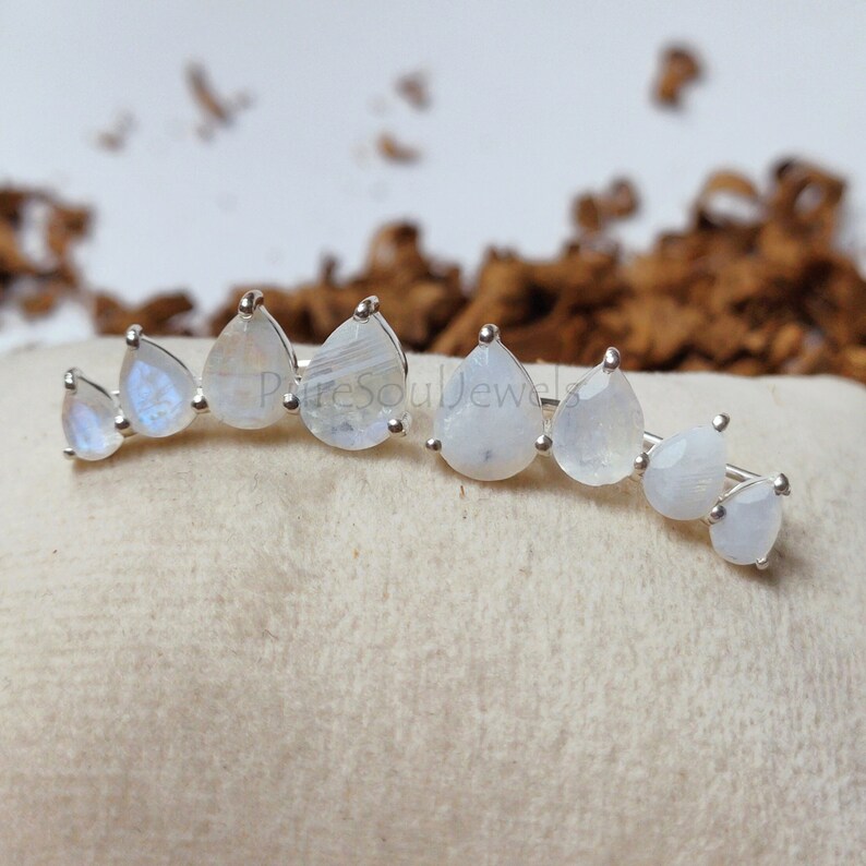May include: A pair of silver ear climbers featuring a row of graduated pear-shaped moonstones. Each earring displays four translucent, iridescent stones set in metal prongs, arranged in a curved line that follows the shape of an earlobe.
