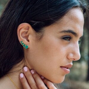 May include: A gold ear climber earring featuring four small, vibrant green teardrop-shaped stones arranged in a graduated line that follows the curve of the earlobe. The metal setting is a polished gold tone, providing a bright contrast.