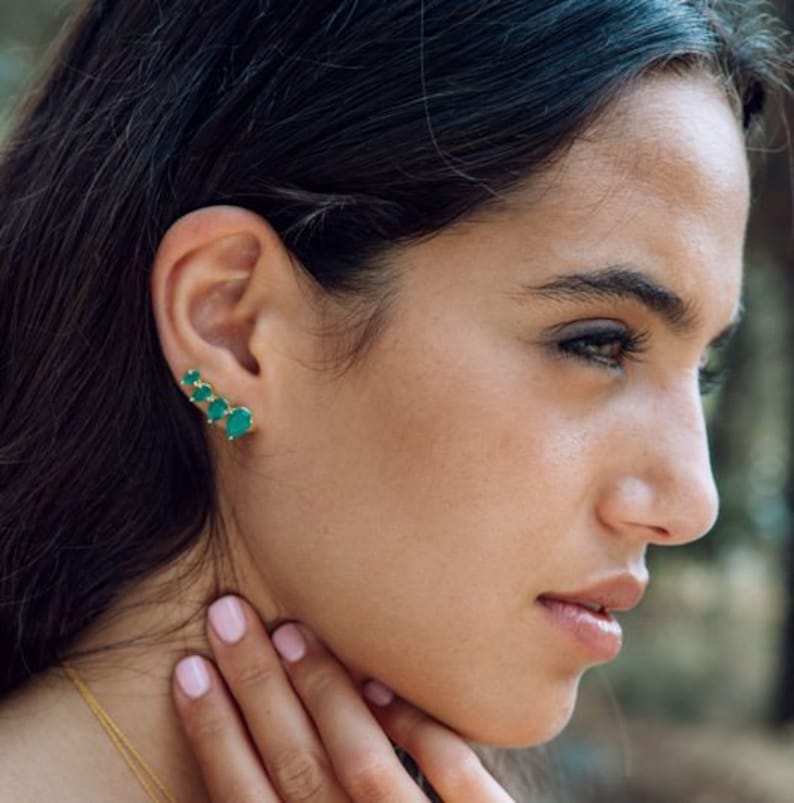 May include: A gold ear climber earring featuring four small, vibrant green teardrop-shaped stones arranged in a graduated line that follows the curve of the earlobe. The metal setting is a polished gold tone, providing a bright contrast.