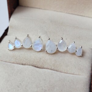 May include: A pair of silver ear climbers featuring a row of four pear-shaped moonstones each, arranged in a graduated size pattern. The stones exhibit a translucent white appearance with subtle blue flashes set in polished silver metal prongs.