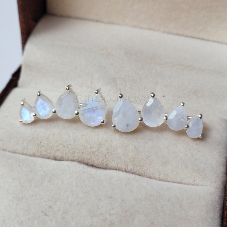 May include: A pair of silver ear climbers featuring a row of four pear-shaped moonstones each, arranged in a graduated size pattern. The stones exhibit a translucent white appearance with subtle blue flashes set in polished silver metal prongs.