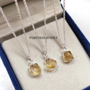 May include: Three silver necklaces with raw, golden-yellow citrine pendants. The pendants are irregularly shaped and set in silver bezels. The necklaces are displayed in a blue jewelry box. The text "PURESOULJEWELS" is visible.