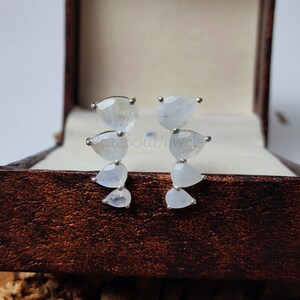 May include: A pair of silver earrings, each featuring four pear-shaped moonstones arranged in a vertical, slightly curved line. The stones exhibit a translucent, milky white appearance with subtle blue iridescence. The text PureSoulJewels is visible.