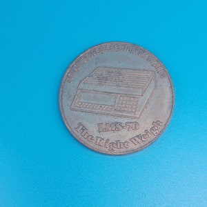 May include: A silver-colored coin with the text "Pitney Bowes Electronic Scales LMS-70 The Right Weigh" and a depiction of a scale.