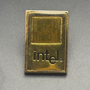 May include: A gold-colored Intel pin with a rectangular shape. The pin features the Intel logo in a stylized font at the bottom and a textured square at the top. The pin is set against a dark gray background.