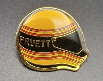 May include: A gold and black enamel pin shaped like a racing helmet with the word "Pruett" on the front.