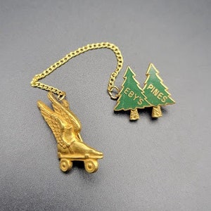 May include: Gold-tone roller skate charm with a green enamel pine tree charm attached to a gold chain. The pine tree charm has the text "Pines Eby's" on it.