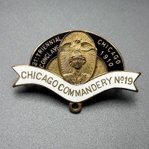 May include: A gold and black enamel pin with a white banner that reads "Chicago Commandery No. 19". The pin features a black and gold design with the text "31st Triennial Conclave 1910 Chicago" and a depiction of a woman with wings.