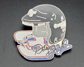 May include: A silver and black pin featuring a race car driver's helmet with the text "Goodyear Service Rd" and a signature that reads "Dale Earnhardt Jr."