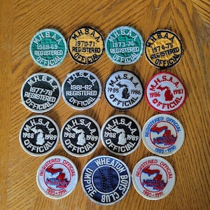 Könnte beinhalten: Eine Sammlung von 12 runden gestickten Aufnähern mit dem Text "MHSAA" und verschiedenen Jahren. Die Aufnäher sind weiß, schwarz, grün, gelb und rot mit blauen und weißen Umrissen des Bundesstaates Michigan. Die Aufnäher sind alle offiziell registriert.