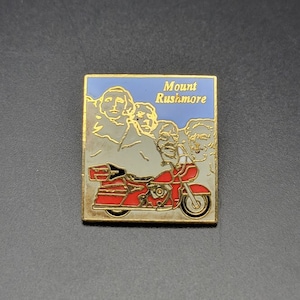 May include: A gold-coloured pin featuring a red motorcycle in front of a blue background with the words "Mount Rushmore" in white. The motorcycle is a Harley Davidson.
