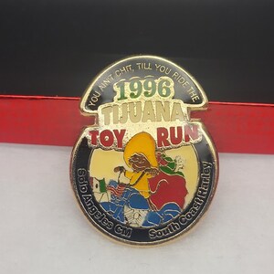 May include: A gold-colored pin with a black background. The pin features a cartoon illustration of a person riding a motorcycle with a bag of toys. The text "YOU AIN'T CHIT, TILL YOU RIDE THE 1996 TIJUANA TOY RUN Solo Angeles CM South Coast Harley" is written around the edge of the pin.