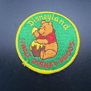 May include: Embroidered patch featuring Winnie the Pooh with a honey pot. The patch is round with a green background and yellow border. Text reads "Disneyland" and "Walt Disney Props".