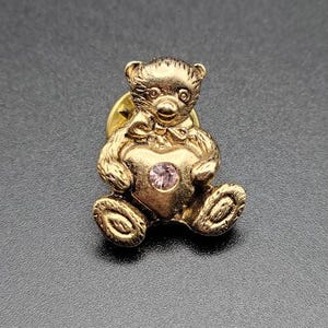 May include: Gold-tone teddy bear pin holding a pink heart with a rhinestone in the center.