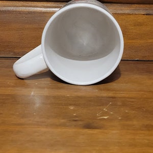 Puede incluir: Taza de caf&eacute; de cer&aacute;mica blanca con asa. La taza est&aacute; vac&iacute;a y se encuentra sobre una superficie de madera.