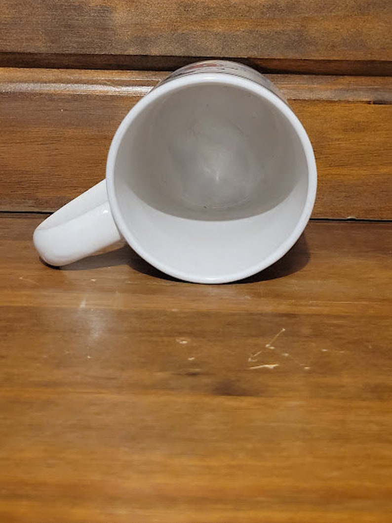 Puede incluir: Taza de caf&eacute; de cer&aacute;mica blanca con asa. La taza est&aacute; vac&iacute;a y se encuentra sobre una superficie de madera.