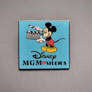 Może przedstawiać: Niebieska i złota przypinka przedstawiająca Myszke Miki trzymającą klapkę z napisem "ACTION". Na przypince widnieje również napis "Disney MGM Studios" w kolorze białym.