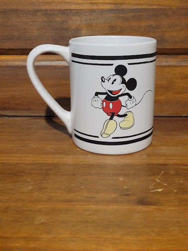 Puede incluir: Taza de cer&aacute;mica blanca con asa negra y rayas negras alrededor de la parte superior e inferior. La taza presenta una imagen de dibujos animados de Mickey Mouse con pantalones cortos rojos y zapatos amarillos caminando.