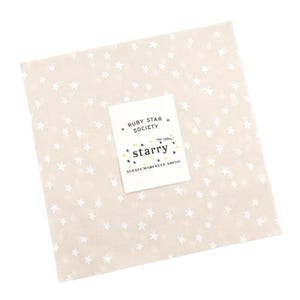 May include: A square, cream-coloured fabric featuring a repeating pattern of white stars. A white label with the text "RUBY STAR SOCIETY" and "starry" is placed on the fabric. The label also includes the text "ALEXIA MARCELLE ABECG".