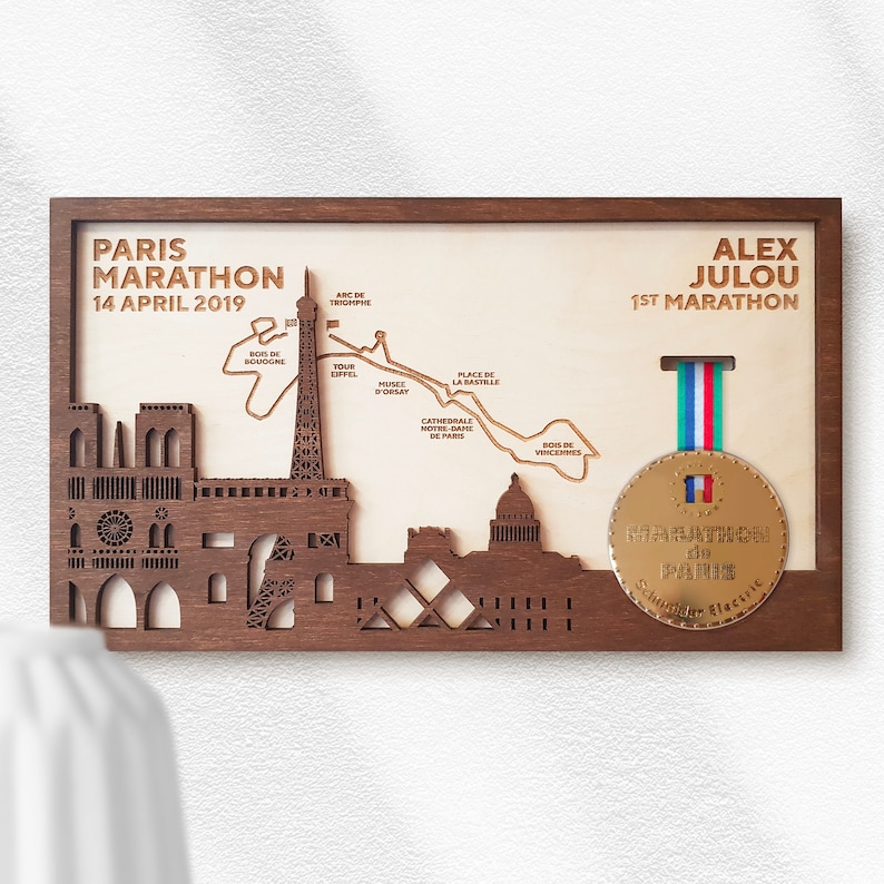 May include: A wooden plaque with a laser-cut design of the Paris skyline, including the Eiffel Tower, Notre Dame Cathedral, and the Arc de Triomphe. The plaque also features a map of the Paris Marathon route and a gold medal with the text "Marathon de Paris". The text "Paris Marathon 14 April 2019" and "Alex Julou 1st Marathon" are also included on the plaque.
