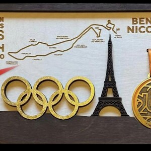 May include: A gold-colored finisher's medal with the text "FINISHER #7195" and a design of the number 2024. The medal is hanging from a brown ribbon with the text "MARATHON POUR TOUS PARIS 2024 AUGUST 10" and a map of the marathon route. The medal is displayed in a wooden frame with a black silhouette of the Eiffel Tower.