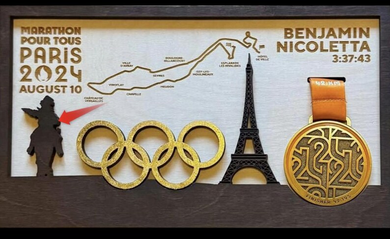 May include: A gold-colored finisher's medal with the text "FINISHER #7195" and a design of the number 2024. The medal is hanging from a brown ribbon with the text "MARATHON POUR TOUS PARIS 2024 AUGUST 10" and a map of the marathon route. The medal is displayed in a wooden frame with a black silhouette of the Eiffel Tower.