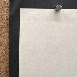Op de afbeelding: Close-up van een prikbord met een zwarte rand en een witte papieren inzet. Een zilveren punaise bevestigt het papier aan het bord. Het papier heeft een textuur.