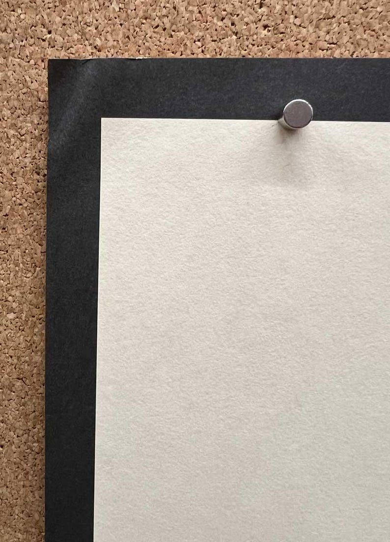 Op de afbeelding: Close-up van een prikbord met een zwarte rand en een witte papieren inzet. Een zilveren punaise bevestigt het papier aan het bord. Het papier heeft een textuur.