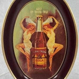 May include: A vintage oval metal tray with a gold rim and a burgundy interior. The tray features a colorful illustration of a Royal Crown Cola bottle with two women in white dresses. The text "Best by Taste-Test" and "Royal Crown RC Cola" is visible on the tray.