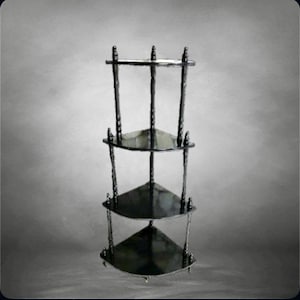 Op de afbeelding: Een donker metalen hoekrek met drie driehoekige niveaus. Het rek heeft een sierlijk ontwerp met verticale steunen en een kleine bovenrail. De planken zijn donker, reflecterend en de algehele stijl is vintage.