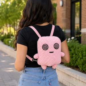 May include: A light pink crocheted backpack shaped like a character with two black spiral eyes and a small black horizontal line for a mouth. The bag features two light pink straps and small protruding arms and legs on the sides and bottom.