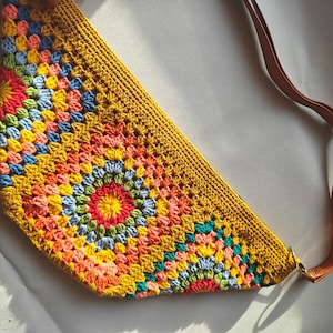 May include: A crocheted bag with a colorful geometric pattern and a brown leather strap. The bag is made with a variety of colors, including yellow, orange, red, green, blue, and pink.