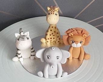 May include: Four small animal figurines made of fondant, including a white zebra with black stripes, a tan giraffe with brown spots, a light brown lion with a textured mane, and a gray elephant, all arranged on a silver circular cake board.