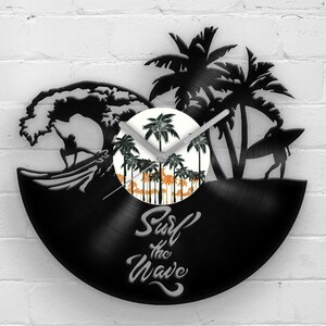 May include: Black vinyl record clock featuring a surfing theme. The clock has a silhouette of a surfer, palm trees, and the words "Surf the Wave." The clock face depicts a sunset scene with palm trees. The clock is mounted on a white brick wall.