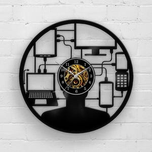 Può includere: Orologio da parete nero con la sagoma di una testa umana, con un quadrante con ingranaggi visibili. Il design comprende contorni di computer e dispositivi mobili collegati alla testa, che simboleggiano la tecnologia e la connettività.