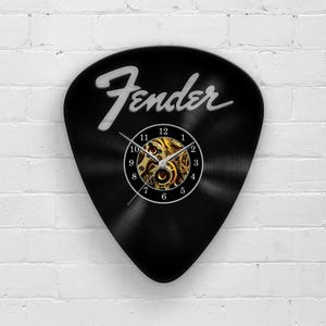 Op de afbeelding: Zwarte gitaar plectrumvormige wandklok met het woord "Fender" in witte letters. De wijzerplaat heeft een zichtbaar goudkleurig mechanisch uurwerk. De wijzers van de klok zijn zwart. De achtergrond is een witte bakstenen muur.