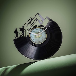 Puede incluir: Un reloj de vinilo negro con un diseño de silueta de montaña. La esfera del reloj presenta un fondo blanco con números y manecillas negros. El reloj está montado sobre una superficie verde.