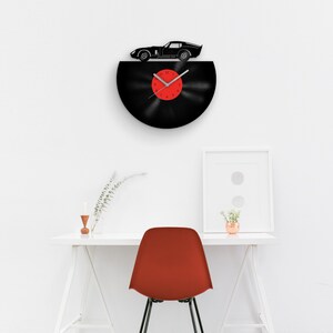 May include: A black vinyl record clock with a red clock face and a black silhouette of a sports car on top. The clock is hanging on a white wall above a white desk with a red chair.