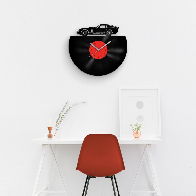 May include: A black vinyl record clock with a red clock face and a black silhouette of a sports car on top. The clock is hanging on a white wall above a white desk with a red chair.