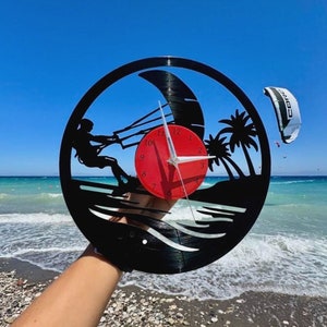 May include: A black vinyl record clock with a red clock face and white hands. The clock is shaped like a wave with a silhouette of a person kitesurfing and palm trees. The clock is being held in front of a beach scene.