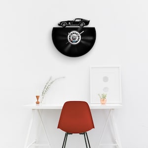 May include: A black vinyl record clock with a black silhouette of a sports car on top. The clock face is a speedometer with a silver needle. The clock is mounted on a white wall above a white desk with a red chair.