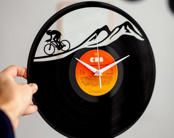 May include: Black vinyl record clock with a cyclist silhouette climbing a mountain. The clock features white hands and a colorful center label. The design includes a mountain range cutout, creating a unique wall decor piece.