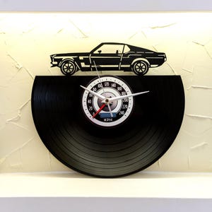 May include: A black vinyl record clock with a classic car silhouette on top. The clock face features a speedometer design with white numbers and hands, and the text "KPH". The clock is mounted on a white surface.