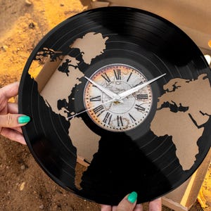 May include: A black vinyl record clock with a world map design. The clock face features Roman numerals and white hands. The map is cut out of the record, revealing a tan background. The clock is held by a person with green nail polish.