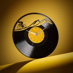 May include: A black vinyl record clock with a yellow face and black hands. The clock features a silhouette of a person riding a bicycle up a mountain.