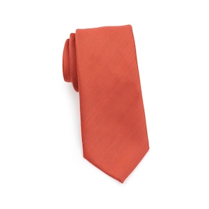 May include: A rolled-up, solid-colored tie in a warm, reddish-orange hue. The tie has a subtle textured pattern and a classic pointed end. The background is plain white, highlighting the tie's color and design.